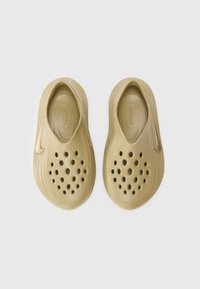 Ljusbruna slip-ons med en texturerad yta, stora cirkulära perforeringar och en glänsande Nike-logotyp som accent på sidan.