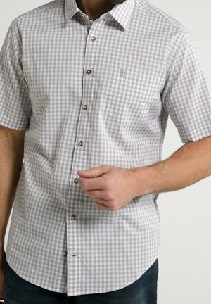Mężczyzna w krótkim rękawie, jasnoszarej i białej w kratę koszuli zapinanej na guziki, z kieszenią na piersi i brązowymi guzikami, zapinający guzik.