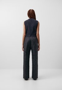 Žena s copánky, oblečená v bezrukávové černé vestě a prošívaných širokých černých kalhotách, stojící na pozadí čistě bílé. Lin