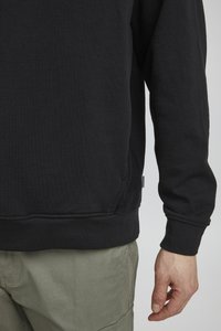 Svart bomullssweatshirt med ribbet rund hals, lange ermer og en subtil sidesømsetikett, kombinert med olivengrønne bukser.