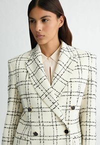 Blazer croisé en tissu texturé crème avec un motif à carreaux noir, doté de quincaillerie dorée et d'un col à revers.