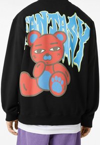 Svart sweatshirt med en färgglad björn grafiskt mönster i rött och blått, med fet text "CANTADY" i grönt och ljusblått på baksidan.