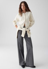 Crème geknoopte cardigan met knopen, gedragen over een zwarte riem, gecombineerd met grijze jeans met een wijde pijp en zwarte enkellaarzen. Gladde textuur en ontspannen pasvorm.