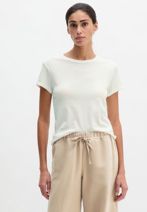 Frau mit zurückgebundenem dunklem Haar, die ein schlichtes weißes T-Shirt mit kurzen Ärmeln und beigefarbene Hosen mit Kordelzug trägt, steht vor einem einfarbigen weißen Hintergrund.