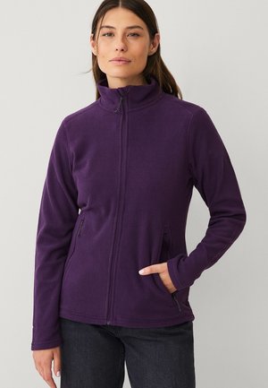 Vrouw met lang bruin haar, gekleed in een paars fleecejack met ritssluiting en een zwarte broek, staand tegen een effen lichte achtergrond.