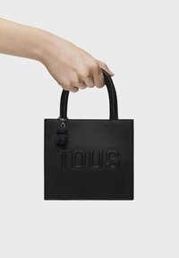 Czarna torba typu tote z teksturowanej skóry z wytłoczonym logo "TOUS", górnymi uchwytami oraz przymocowanym czarnym charmem.