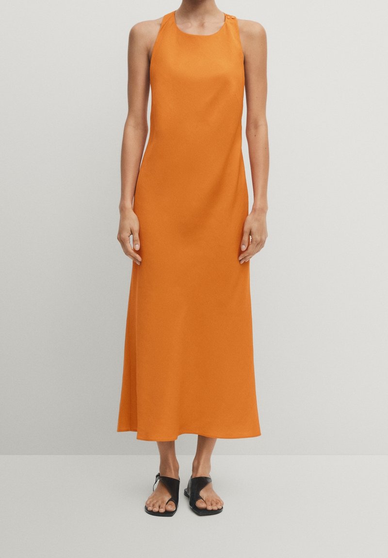 Robe midi orange sans manches avec détail boutonné à l'épaule, portée avec des sandales plates noires à bout ouvert sur un fond uni.