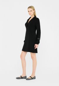 Abito nero con scollatura a V, maniche lunghe e design aderente. Abbinato a ballerine con motivo bianco e nero, dotate di una fascia e dettagli decorativi.
