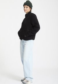 Giacca in pile nera con chiusura a zip, colletto rialzato e tasche frontali. Abbinata a jeans a gamba larga di colore azzurro chiaro e sneakers grigie.