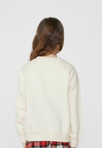 Felpa color crema con scollatura rotonda, spalle ribassate e polsini a coste, indossata sopra una gonna a quadri rossa e nera.