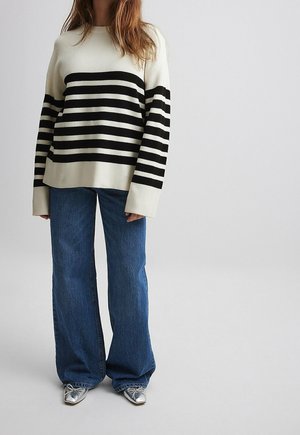 Personne portant un pull blanc à rayures noires horizontales, un jean bleu à jambes larges et des chaussures argentées à bouts pointus, debout devant un fond uni.