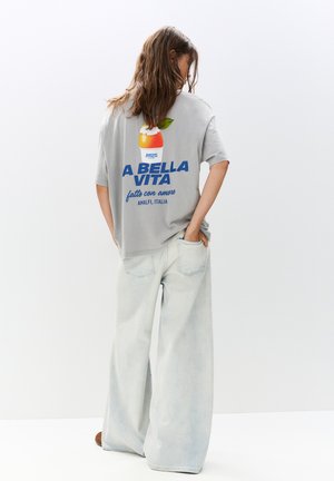 Tricou oversized gri, cu un grafic colorat de fructe și text pe spate, purtat cu blugi evazați de un albastru deschis. Material moale din bumbac.
