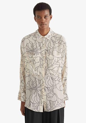 Persona che indossa una camicia a maniche lunghe con bottoni con motivi floreali astratti a linee nere su tessuto panna e pantaloni neri.