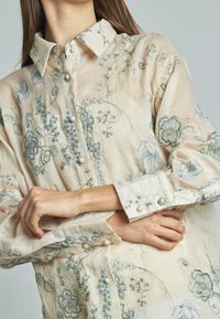 Camicia button-up di un beige chiaro con ricami floreali verdi e blu. Tessuto morbido e strutturato con colletto classico e bottoni perla.