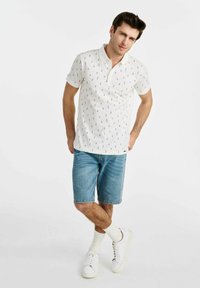 Biała koszulka polo z małym czarnym wzorem, w zestawie z jasnymi dżinsowymi szortami. Model nosi białe trampki i białe skarpetki typu crew.