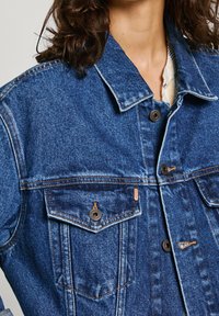 Jeansjacka i blått med krage, två bröstfickor, knäppningar med knappar och kontrasterande sömmar. Tyget har en texturerad, urblekt look.