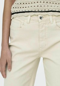 Los pantalones vaqueros de mezclilla beige presentan un corte ajustado, con un cierre de botón único y dos bolsillos frontales. La tela tiene una textura suave y costuras mínimas.