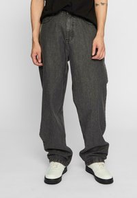 Mørkegrå, vide jeans med en løs pasform, lavet af denim. Har klassiske lommer og en simpel lukning i taljen. Skoene er hvide med sorte såler.