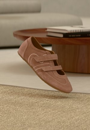 Pantof maro din piele întoarsă cu două curele Velcro, așezat pe un covor bej țesut, în fața unei măsuțe de lemn cu cărți așezate una peste alta.