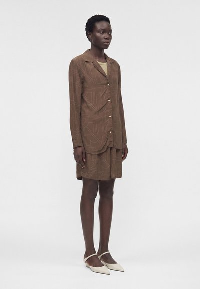 Γυναίκα που φοράει ένα καφέ πουκάμισο με κουμπιά και μακριά μανίκια με σχέδιο και ταιριαστά σορτς, καθώς και μπεζ μυτερά φλατ παπούτσια, στέκεται απέναντι από ένα απλό φόντο.