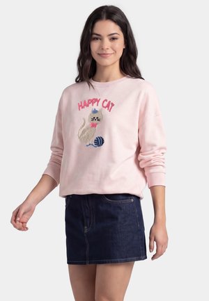 Kvinde med langt mørkt hår iført en lyserød sweatshirt med en kat og garn-grafik og ordene "Happy Cat,", kombineret med et mørkt denimskørt.