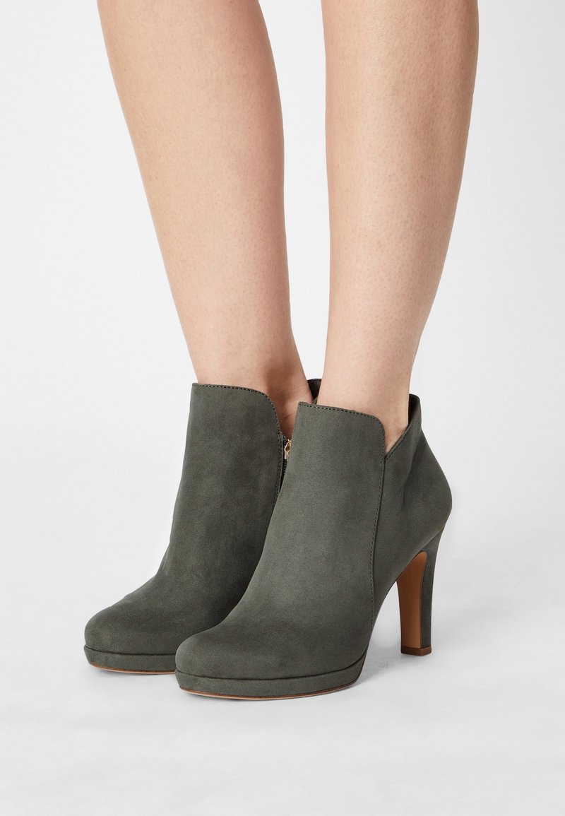 Bottes à talons en daim vert olive montant à la cheville avec fermetures éclair latérales, portées sur des jambes nues sur un fond blanc uni.