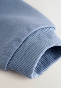 Vaaleansininen collegepaita, jonka hihassa on sileä, pehmeä tekstuuri ja ribannut päällinen. Kangas näyttää joustavalta ja hyvin viimeistellyltä.