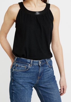 Frau trägt ärmelloses schwarzes Oberteil mit weitem, geripptem Ausschnitt und blaue Jeans, Hand auf der Hüfte abgestützt.