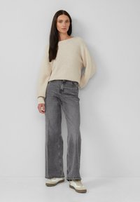 Donna con lunghi capelli scuri che indossa un maglione in maglia color crema, pantaloni a vita alta grigi sbiaditi e scarpe da ginnastica bianche, in piedi contro un muro semplice.