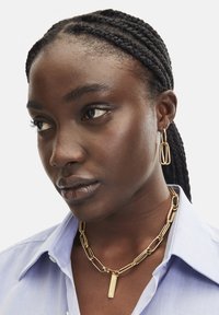 Goldkette mit verlängerten Gliedern und einem rechteckigen Anhänger; kombiniert mit goldenen Creolen. Das Modell trägt ein hellblaues Hemd mit Knöpfen.