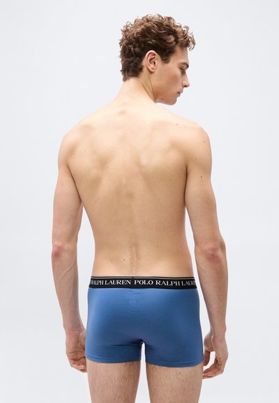 Νεαρός άνδρας με σγουρά μαλλιά, γυμνός από τη μέση και πάνω, γυρισμένος πλάτη, φοράει μπλε ανδρικά μποξεράκια Polo Ralph Lauren.