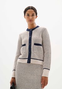 Knit cardigan in cream with navy trim, featuring a round neckline, five buttons, and two front pockets with a textured fabric.