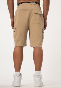 Shorts cargo beiges avec poches latérales, taille élastique et détails de logo. Présente une coupe décontractée, atteignant la longueur mi-cuisse.