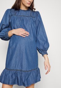 Τζιν φόρεμα με φουσκωτό λαιμό, φουσκωτά μανίκια και ρεβέρ στο τελείωμα. Το ύφασμα είναι σε μέτρια μπλε απόχρωση με μαλακή υφή.