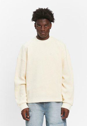 Cream-colored textured sweatshirt with a round neckline, dropped shoulders, and ribbed cuffs, worn with light blue cargo jeans.