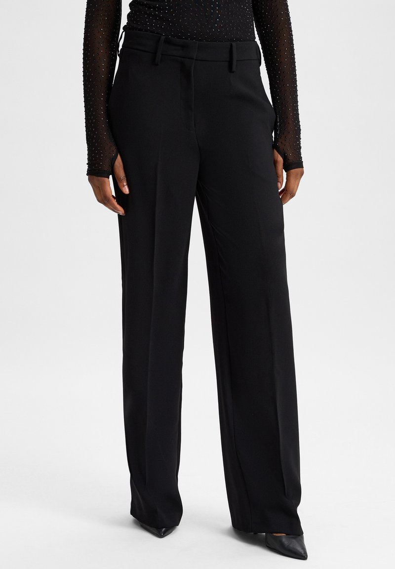 Zwarte wijde broek met een gladde textuur, voorzien van een taillerand en subtiele riemlussen. Gecombineerd met puntige zwarte schoenen.