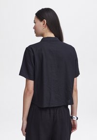 Camicia nera in lino tagliata con maniche corte, colletto su misura e superficie leggermente testurizzata. Indossata con pantaloni neri semplici e bracciale argentato.