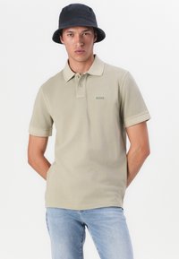Beige polo skjorte med kragehals, korte ermer og grønt logo. Brukt sammen med blå jeans og en svart bøttehatt.
