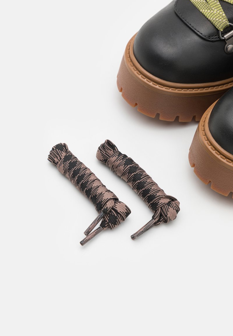 Stivali in pelle nera con una suola spessa e strutturata di colore marrone. Due paia di stringhe scure, marroni e nere con motivi giacciono accanto a loro.