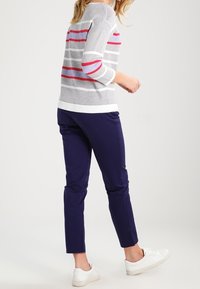 Žena v pruhovaném bílém, šedém a červeném svetru s námořnickými kalhotami a bílými teniskami, stojící bokem na jednobarevném pozadí.