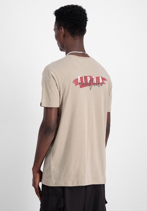 Beige T-Shirt mit kurzen Ärmeln, auf dessen oberem Rücken ein rotes und schwarzes "ALPHA" Grafikdesign zu sehen ist. Hergestellt aus einem glatten Baumwollmischgewebe.