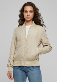 Veste bomber beige clair avec une fermeture éclair frontale, poignets et ourlet côtelés, fabriquée en tissu lisse, dotée de poches latérales et d'un col rond simple.