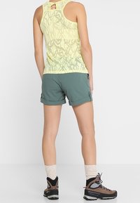 Ljust gult, mönstrat linne med en nyckelhålsrygg, kombinerat med gröna shorts. Bär beige strumpor och bruna vandringsskor med svarta accenter.