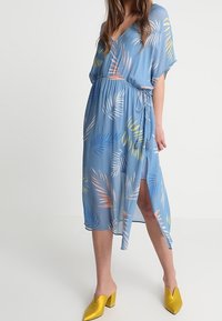 Mujer vestida con un vestido envolvente azul de estampado de hojas, con una abertura frontal y zapatos de tacón amarillo, de pie contra un fondo liso.