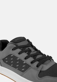 Grauer Sportschuh mit einem strukturierten schwarzen Mesh-Panel, schwarzen Schnürsenkeln und einer Gummisohle. Verfügt über ein Logo auf der Zunge und einen gepolsterten Kragen.