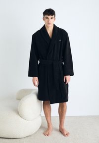Melna halāta ar šalu apkakli, jostas saišu un teksturētu audumu. Modelis stāv basām kājām blakus apaļai krēslā krēmkrāsā.