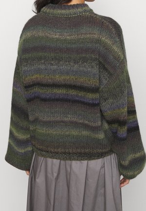 Femme portant un pull en tricot rayé multicolore aux tons verts, violets et gris, associé à une jupe grise, vue de dos.