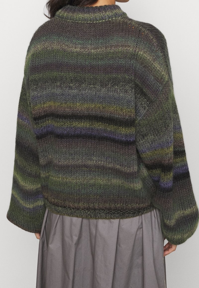 Femme portant un pull en tricot rayé multicolore aux tons verts, violets et gris, associé à une jupe grise, vue de dos.