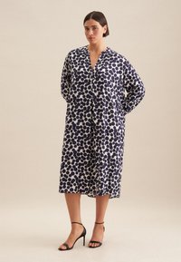 Abito a pois blu navy e bianchi con una vestibilità ampia, senza colletto e maniche lunghe, che arriva a metà polpaccio. Indossato con tacchi neri a strappo.