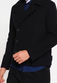 Schwarzer Woll-Pea-Coat mit doppelreihigem Design, großen Revers, zwei Vorderentaschen und glatter Textur. Über einem blauen Hemd getragen.
